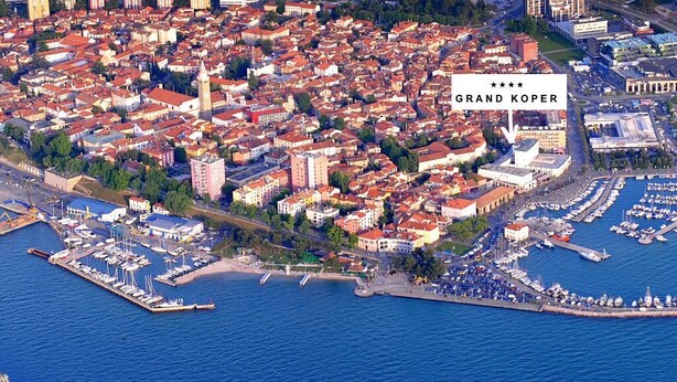 Vista aérea del Grand Kopper, que muestra un paisaje urbano costero con edificios de techo rojo y una marina con barcos, además de un gran crucero amarrado cerca del paseo marítimo.