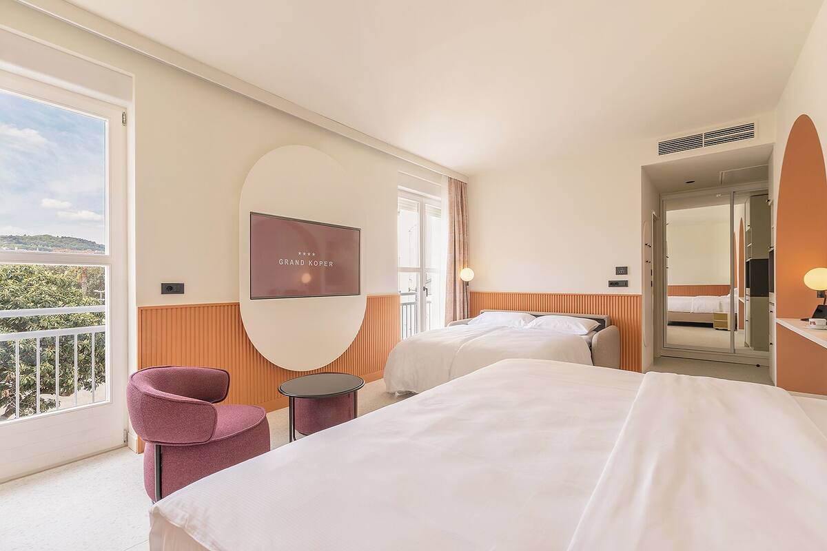 Habitación de hotel con dos camas, televisión, una silla, una mesita y balcón con vistas.