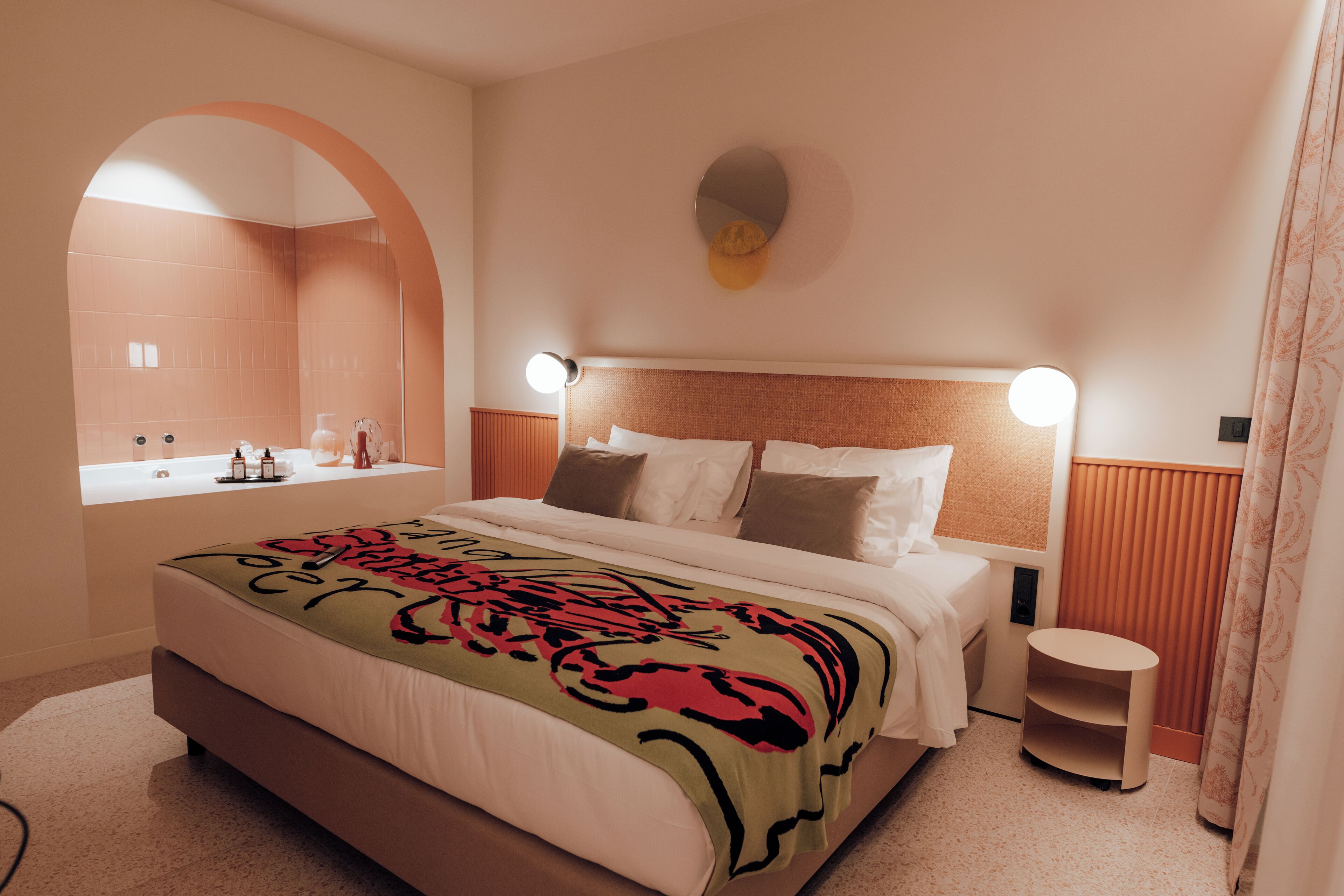 Une chambre d’hôtel moderne avec un grand lit, un miroir rond, une petite table et une fenêtre donnant sur la salle de bain.