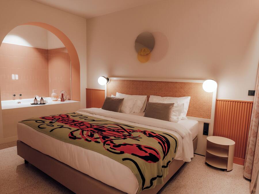 A modern hotel room with a large bed, a round mirror, a small table, and a window with a view of the bathroom.