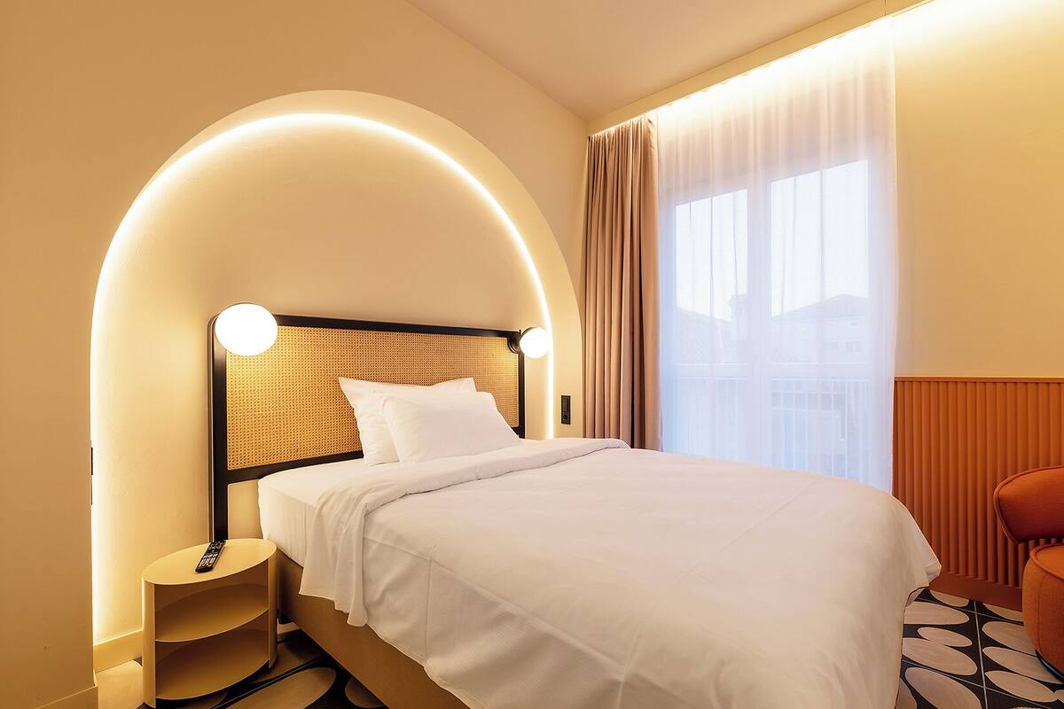 Habitación de hotel con cama, mesita de noche, control remoto y ventana con cortinas.
