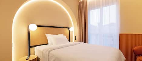 A hotel room with a bed, a bedside table, a remote control, and a window with curtains.