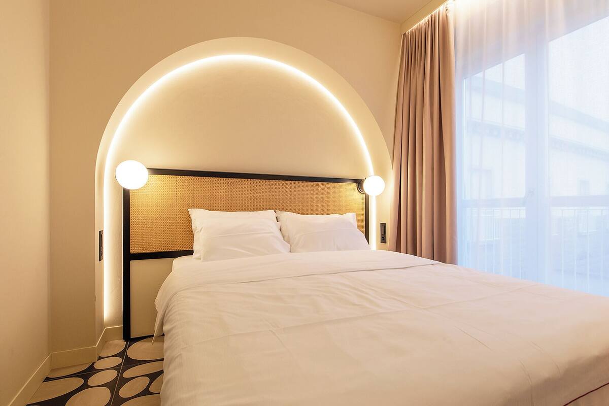 Habitación de hotel con cabecera curva, cama con ropa blanca y vistas al exterior a través de amplios ventanales.