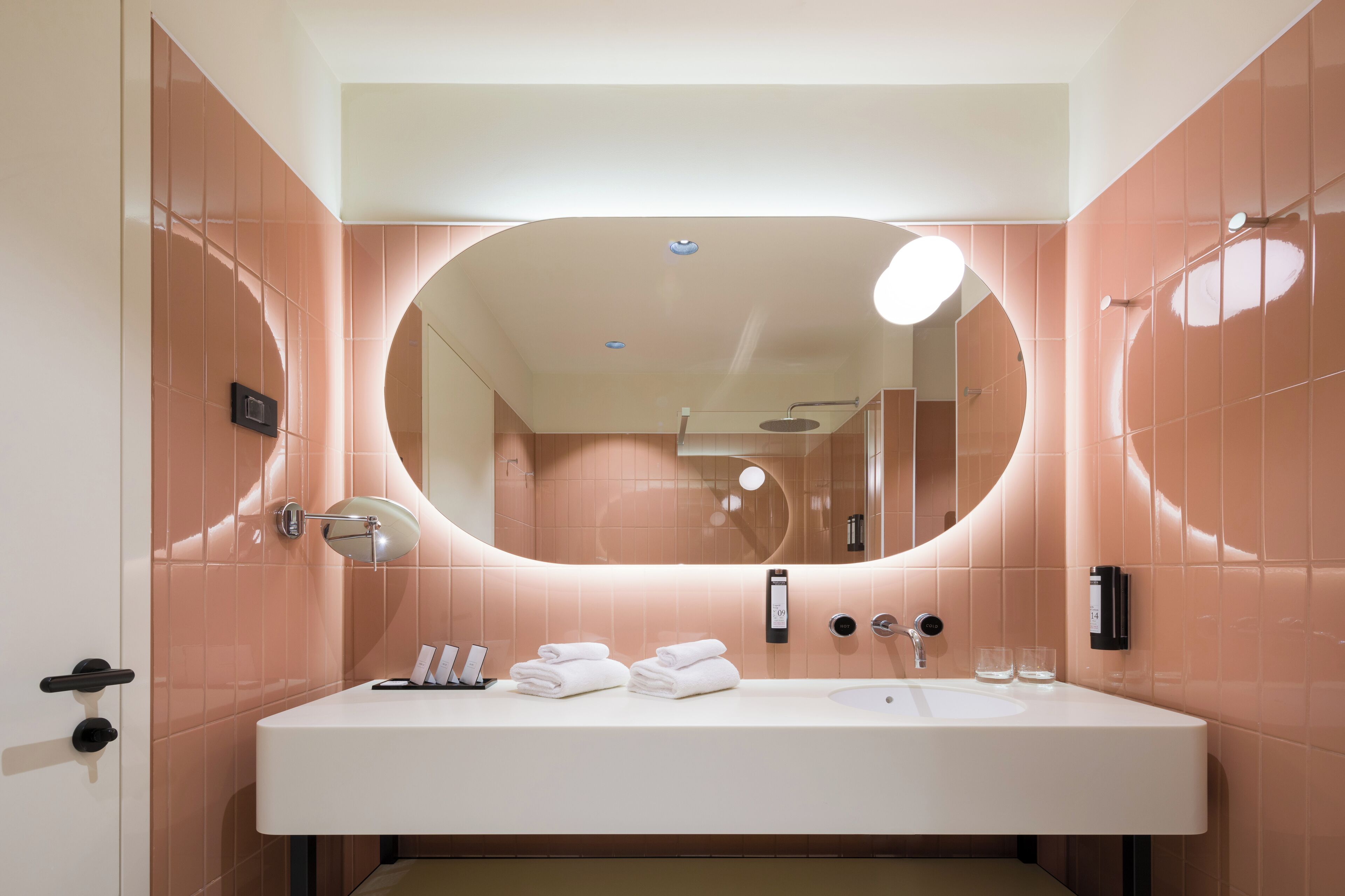 Un baño moderno con un espejo ovalado grande, un lavamanos blanco y paredes con azulejos rosa.