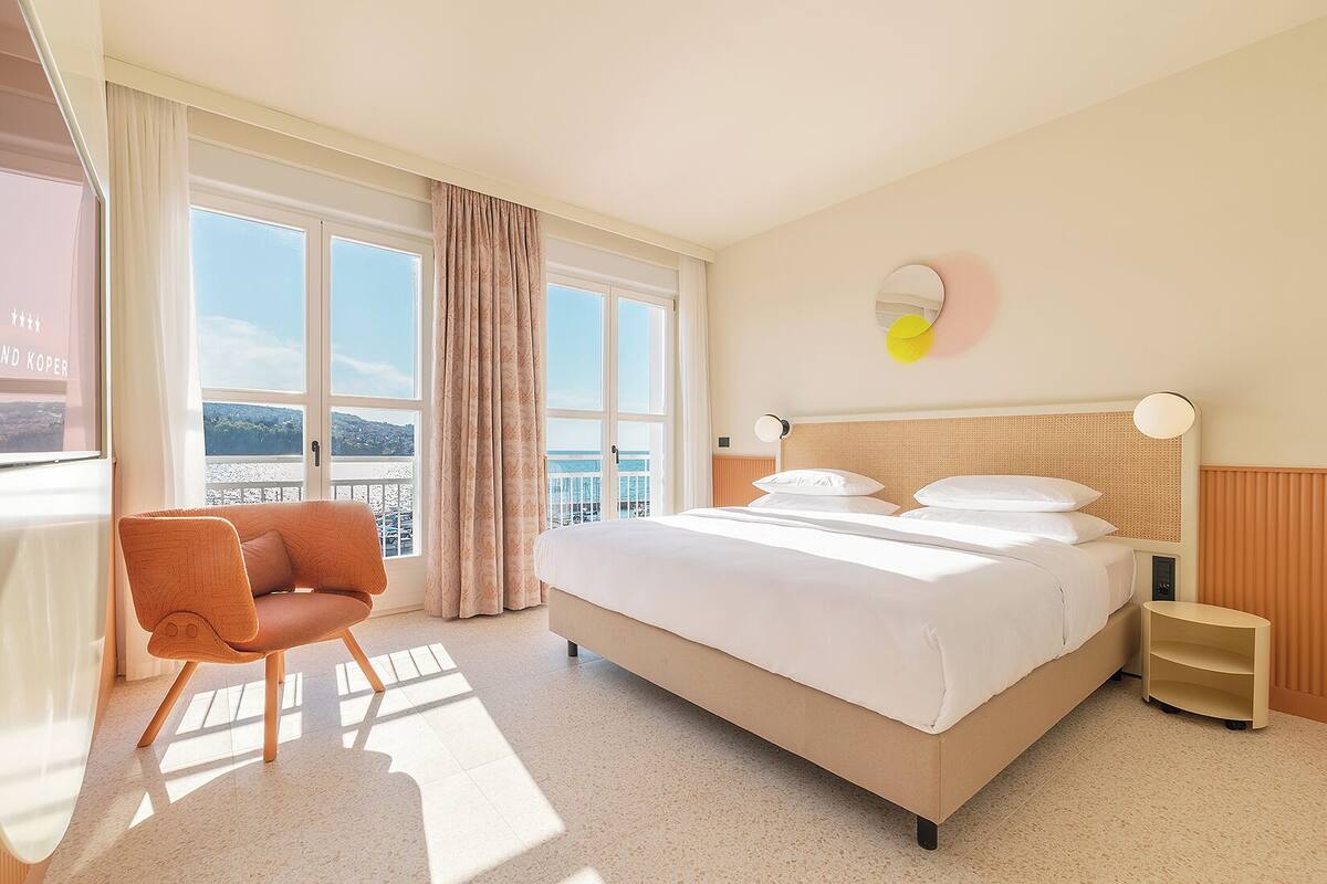 Habitación de hotel con una cama grande, una silla, una mesita de noche y vistas al mar a través de amplios ventanales.