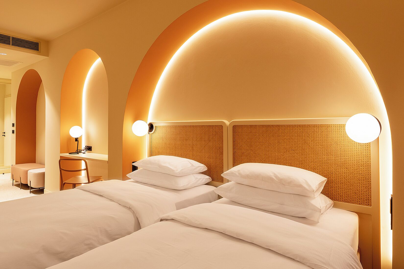 A hotel room with two beds, each with a headboard featuring a woven texture and round wall-mounted lights.