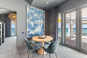 Une chambre d’hôtel moderne avec une table ronde en bois, des chaises à revêtement gris et une vue sur l’océan grâce à de grandes portes coulissantes.