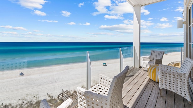 A beach view from a covered deck with white wicker chairs and a yellow cushion.