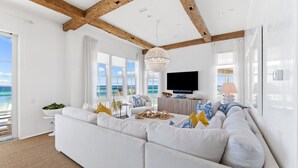 A modern living room with a large white sectional sofa, wooden beams on the ceiling, a chandelier, and a view of the ocean through large windows.