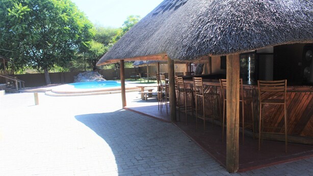 Zona junto a la piscina con techo de paja, mesas y sillas de madera, y vista al estanque.