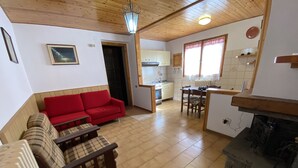 A cozy living area with a red sofa, a plaid sofa, and a wooden table. There is a kitchen with a stove, sink, and window with curtains.