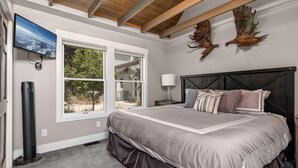Quarto com uma cama grande, televisão na parede, janela com vista para o verde e teto com vigas aparentes.