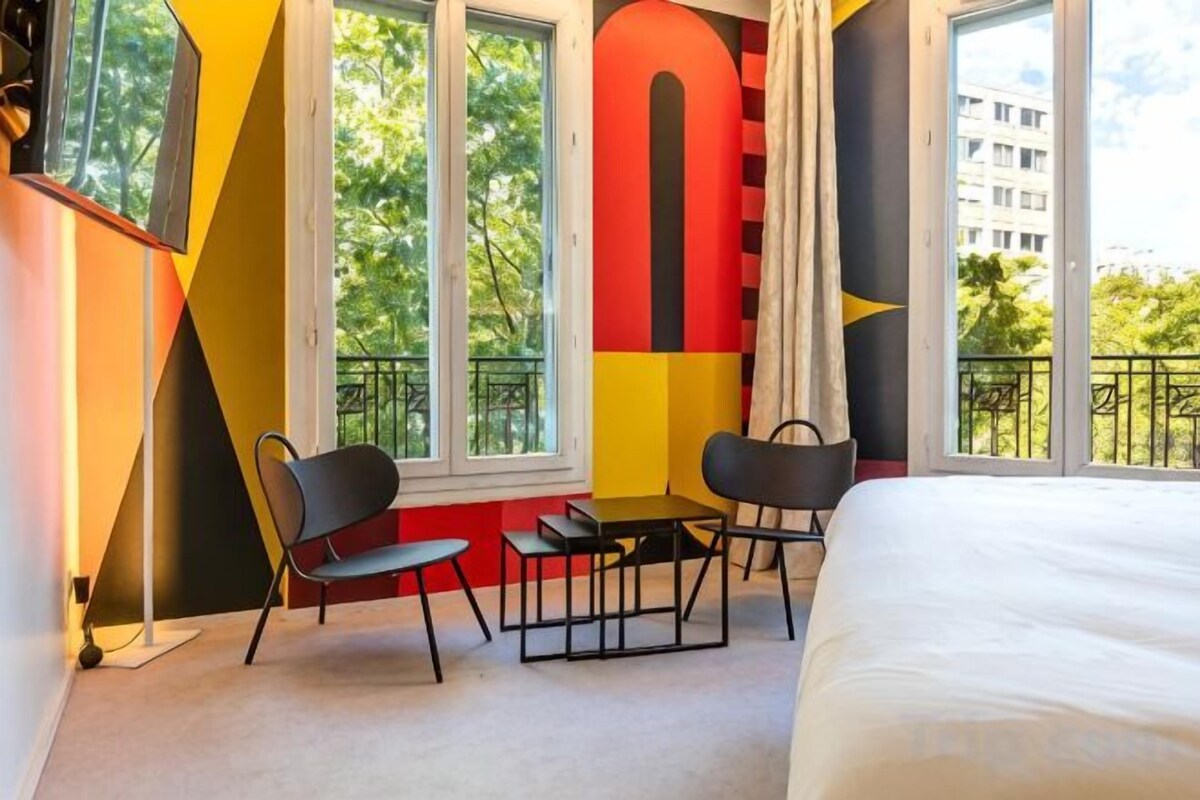 Une chambre d’hôtel avec un lit, deux chaises, une petite table et une grande fenêtre donnant sur l’extérieur.
