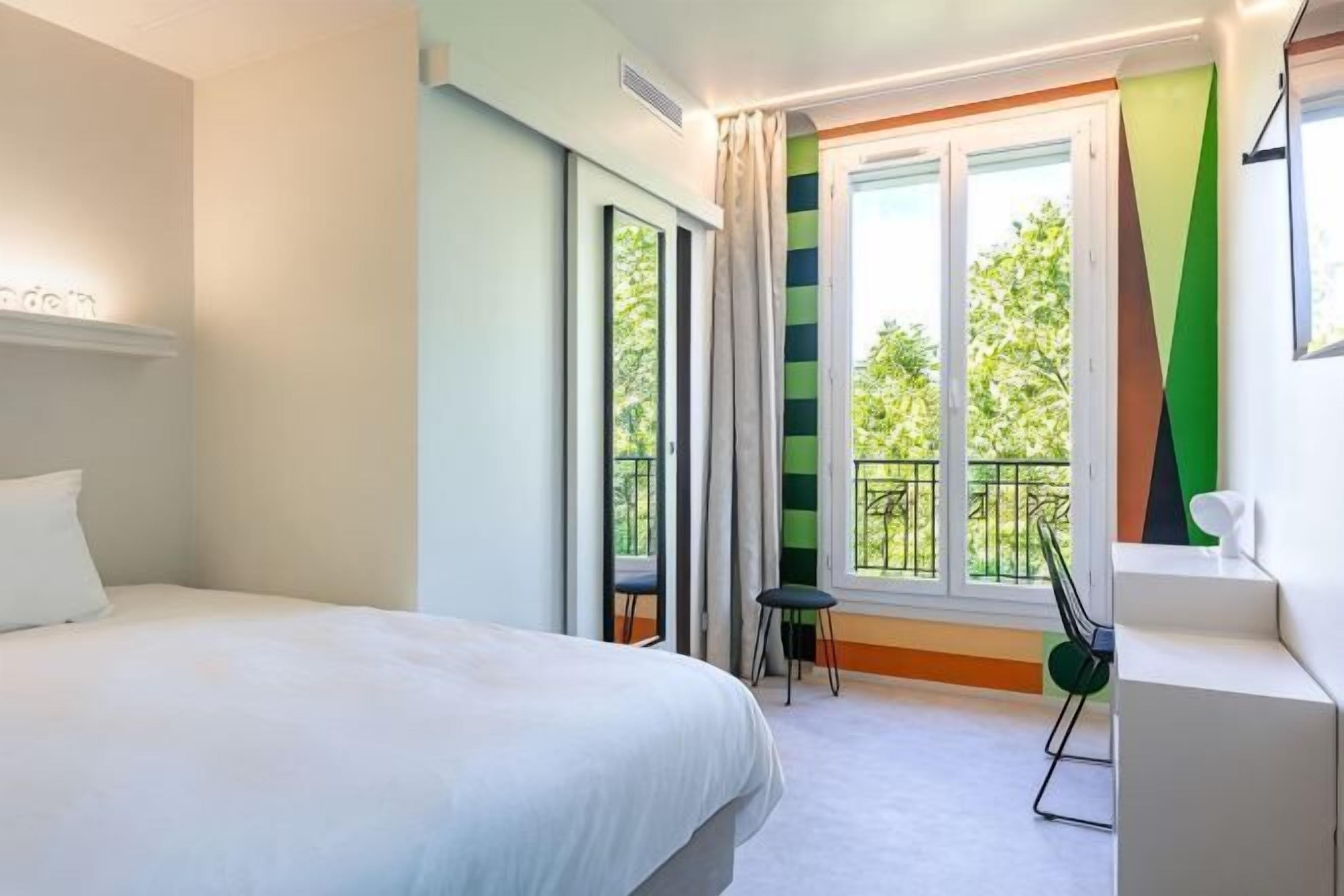 Une chambre d’hôtel comprenant un lit, un bureau, une chaise, une fenêtre avec des rideaux et un balcon.