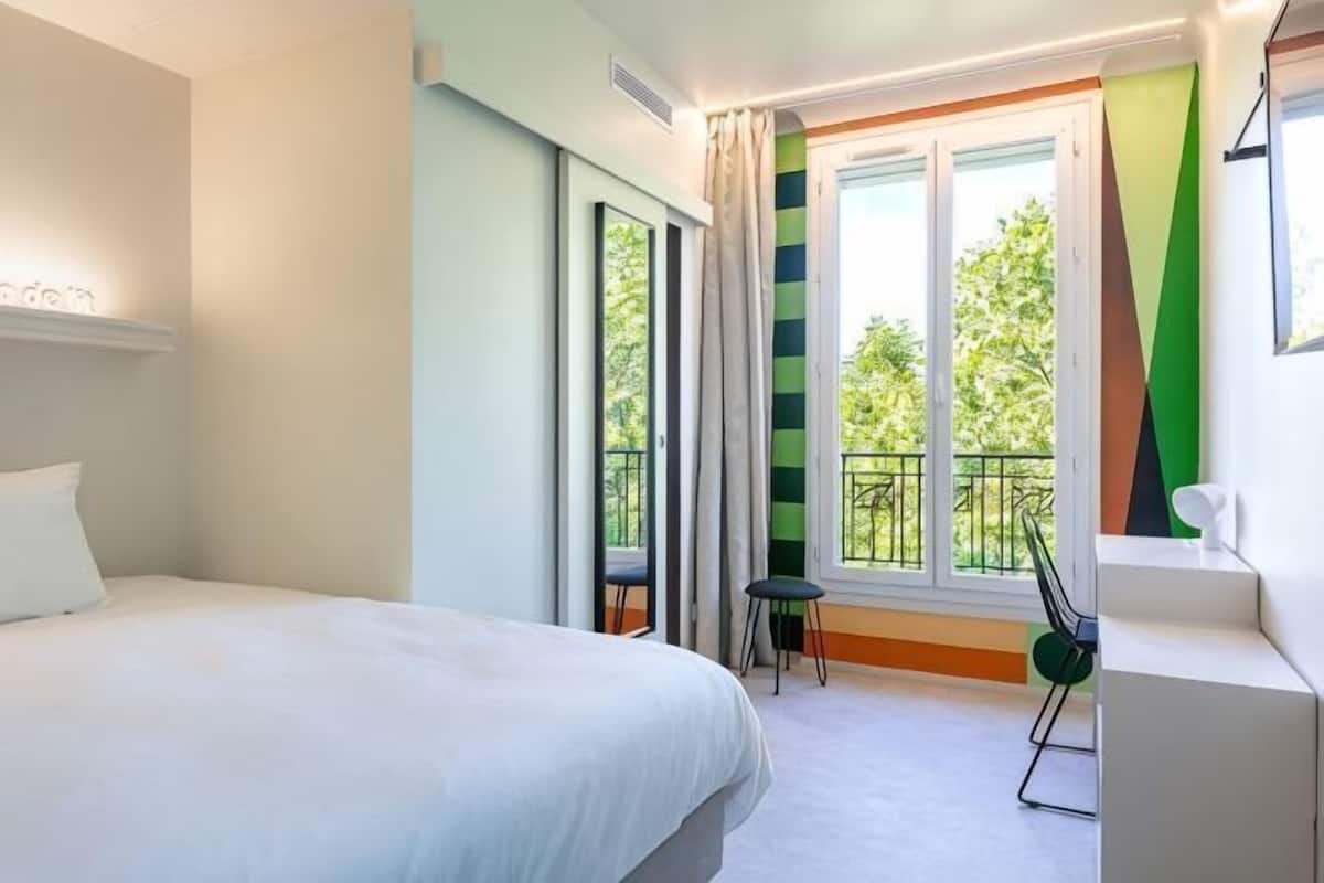 Une chambre d’hôtel comprenant un lit, un bureau, une chaise, une fenêtre avec des rideaux et un balcon.
