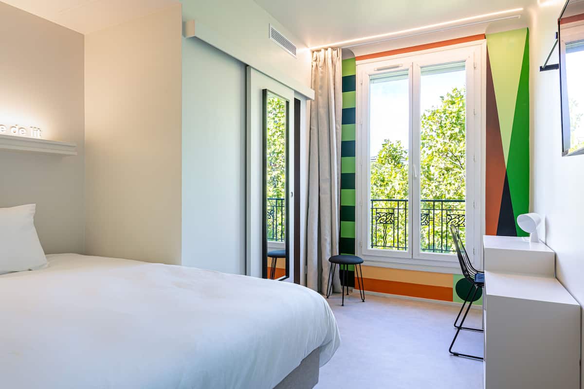 Une chambre d’hôtel moderne, dotée d’un grand lit, d’un bureau et offrant une vue sur l’extérieur grâce à une fenêtre agrémentée de rideaux.