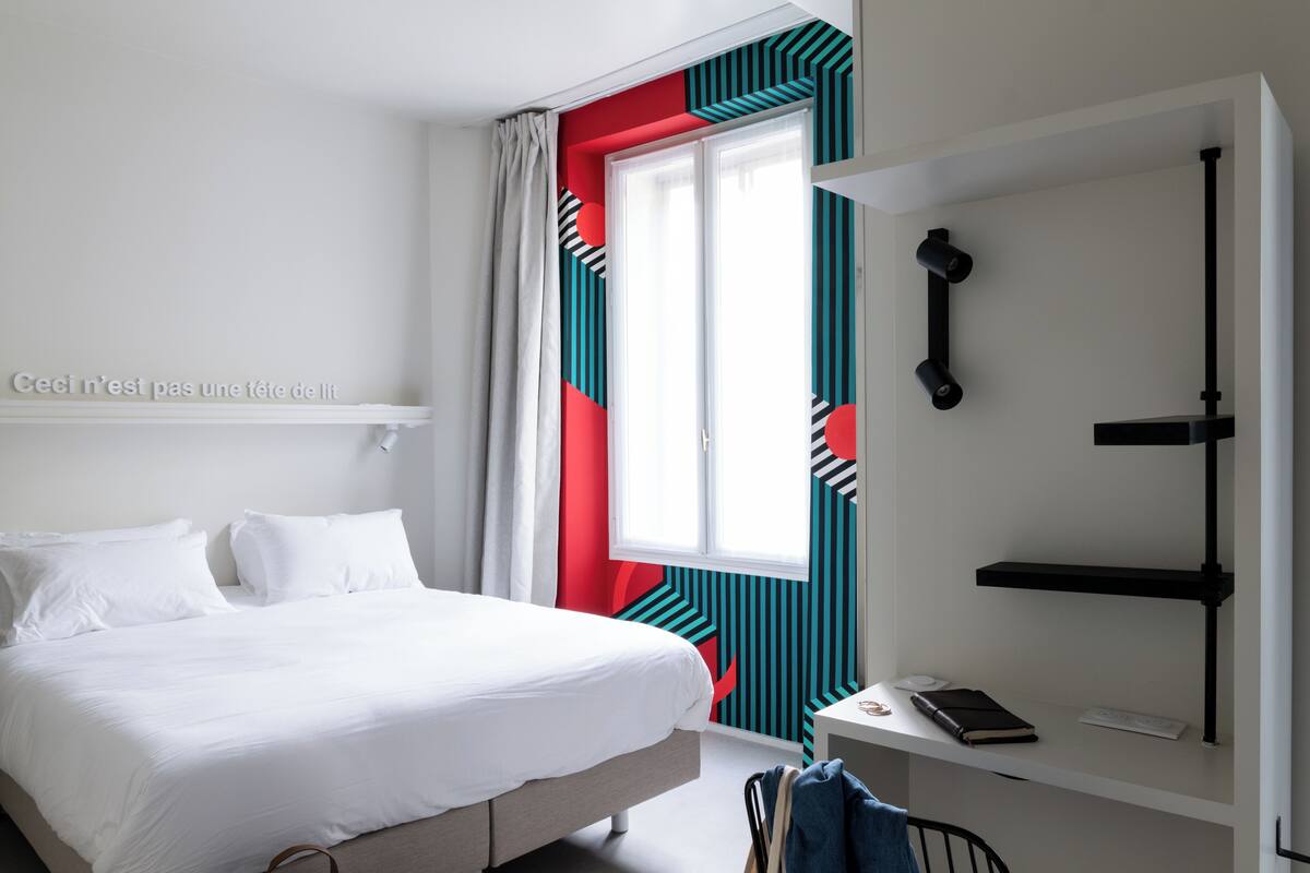 Une chambre à coucher comprenant un lit, un bureau avec une chaise, une fenêtre avec des rideaux et une étagère fixée au mur.