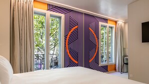A modern hotel room with a large bed, two windows with curtains, a view of trees, and a geometric wall design. - Graphik Montparnasse (Paris)