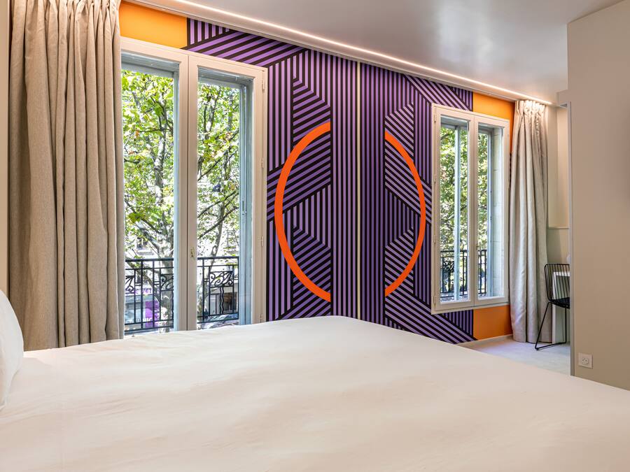 Une chambre d’hôtel avec un grand lit, deux fenêtres avec des rideaux, un mur orné d’un motif géométrique et une vue sur l’extérieur.
