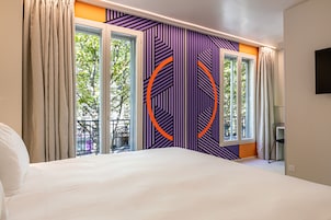 Habitación de hotel con una cama grande, dos ventanas con cortinas, una pared con diseño geométrico y vistas al exterior.