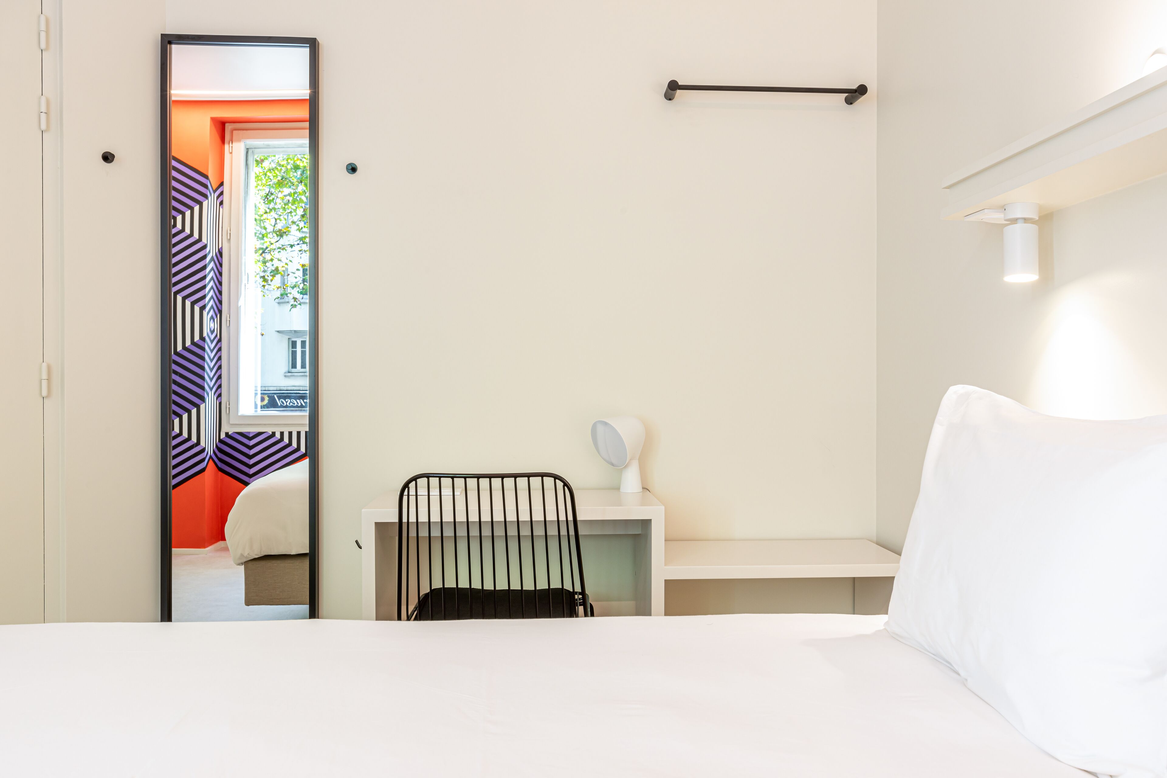 Une chambre d’hôtel comprenant un lit, un bureau avec une chaise, un miroir et une fenêtre donnant sur un immeuble.