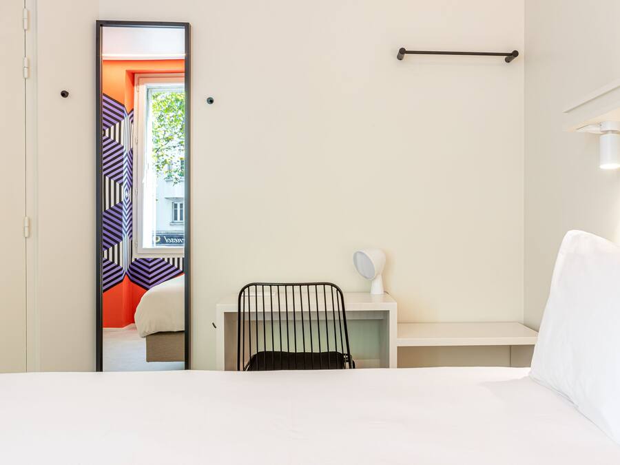 Une chambre d’hôtel comprenant un lit, un bureau avec une chaise, un miroir et une fenêtre donnant sur un immeuble.