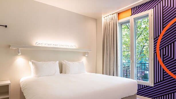 A modern bedroom with a large bed, a headboard with a light fixture, a window with curtains, and a wall with a geometric pattern. - Graphik Montparnasse (Paris)
