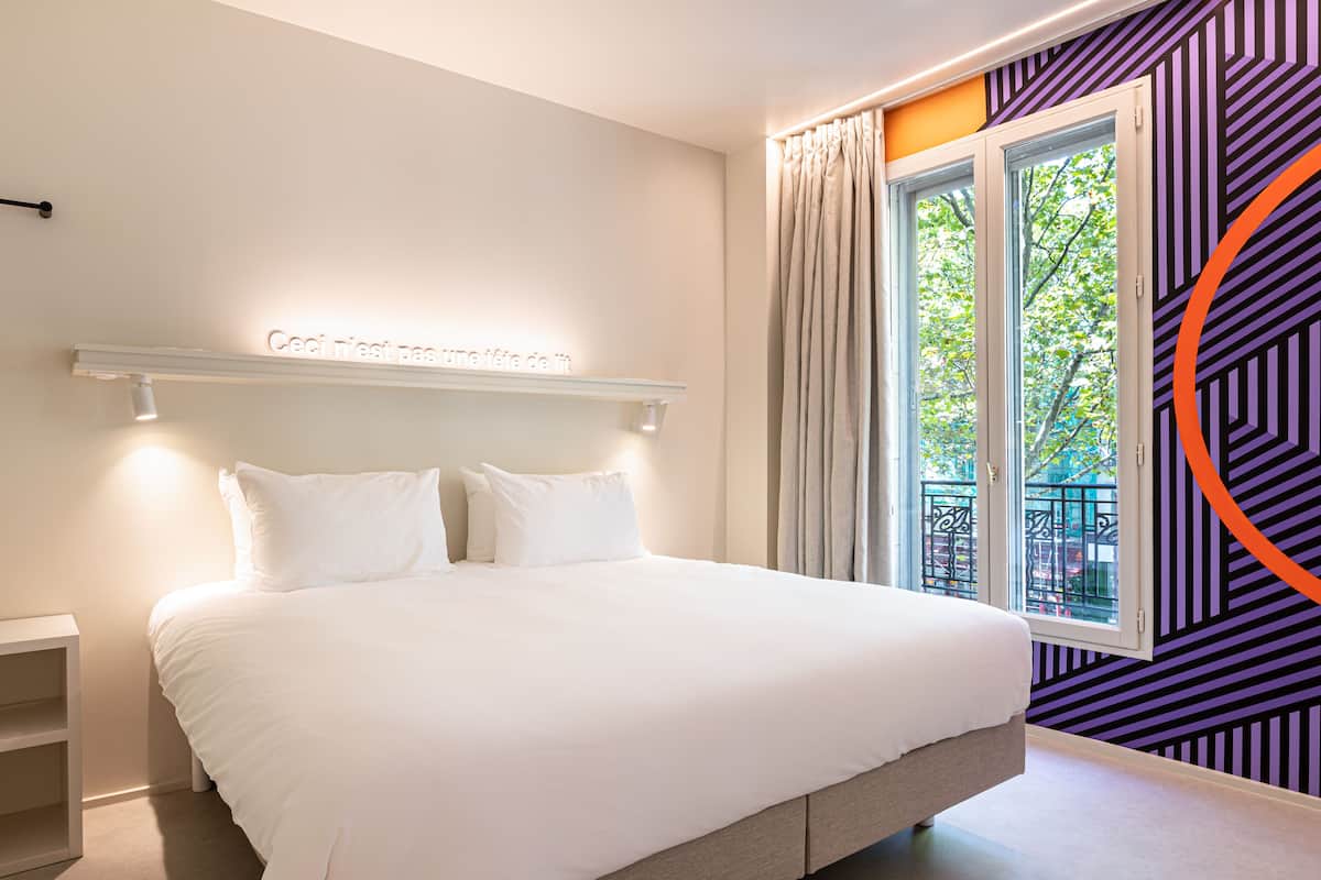 Une chambre moderne avec un grand lit, une table de chevet, une étagère fixée au mur et une fenêtre avec des rideaux.