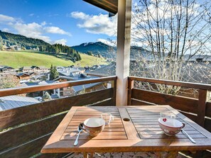 A wooden deck with two tables, each with a bowl and a spoon. A clear glass is also visible. The deck overlooks a mountainous landscape with buildings and trees.