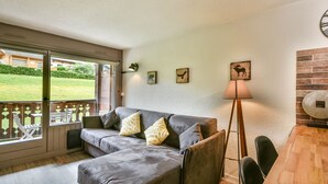 A cozy living room with a grey sofa, a wooden dining table, and a lamp. There are three wall-mounted pictures and a sliding glass door leading to a balcony. - Renovated Studio with Cabin in Village Center, Near Ski Lifts - Les Gets (Les Gets)