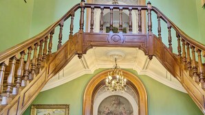 Un magnifique escalier orné de boiseries sculptées et surmonté d’un lustre, menant à une pièce où se trouvent une table à manger et des chaises.
