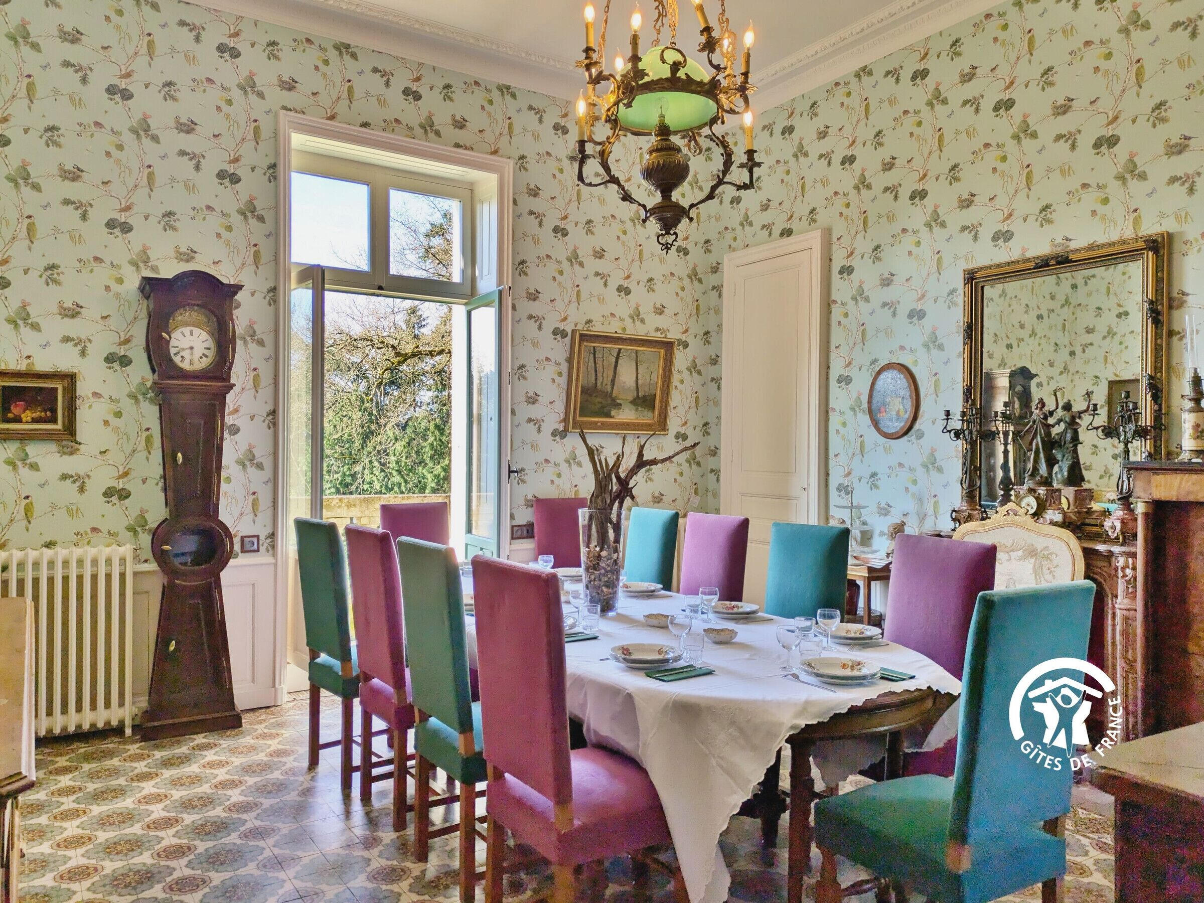 A dining room with a table set for four, surrounded by colorful chairs, a large clock, and a chandelier.