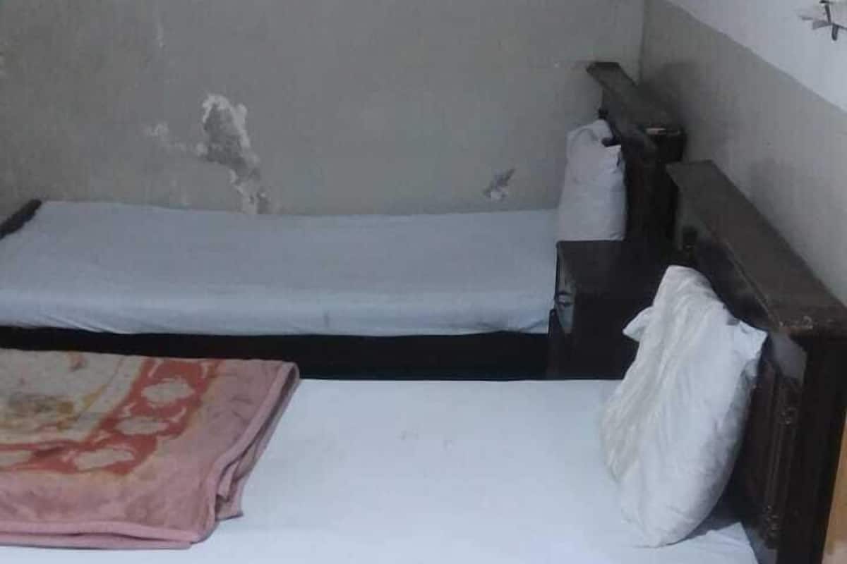 A small, dimly lit room with two single beds, each with a wooden headboard and a white mattress. The room has a tiled floor and a plain, unadorned wall.