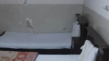 A room with two single beds, each with a pillow and a blanket. The room has a tiled floor and a plain wall with a visible crack.