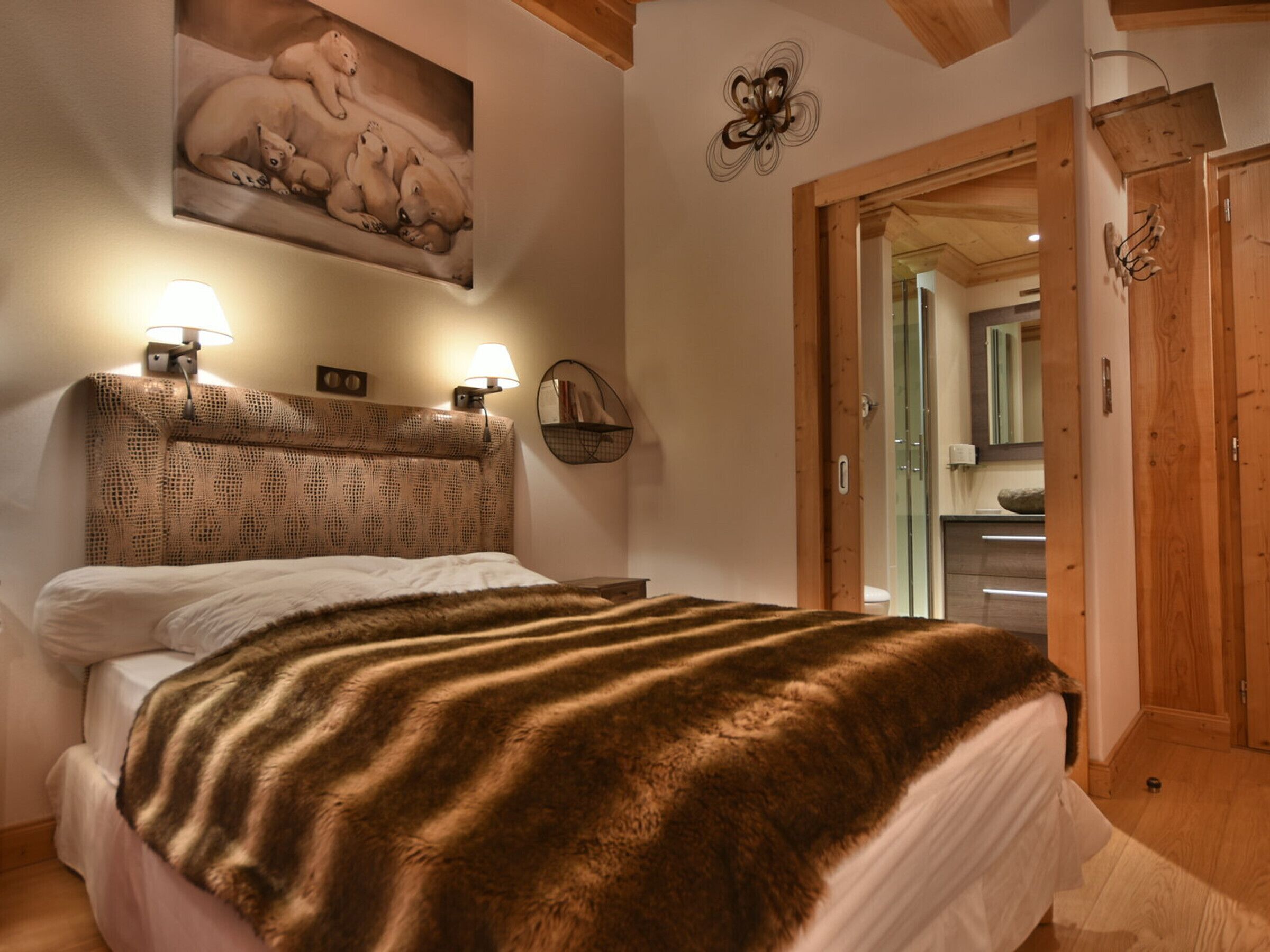 Una camera da letto con un letto grande, una testiera con motivo decorativo, due lampade a parete, un quadro con orsi polari e una porta in legno che dà su un bagno.