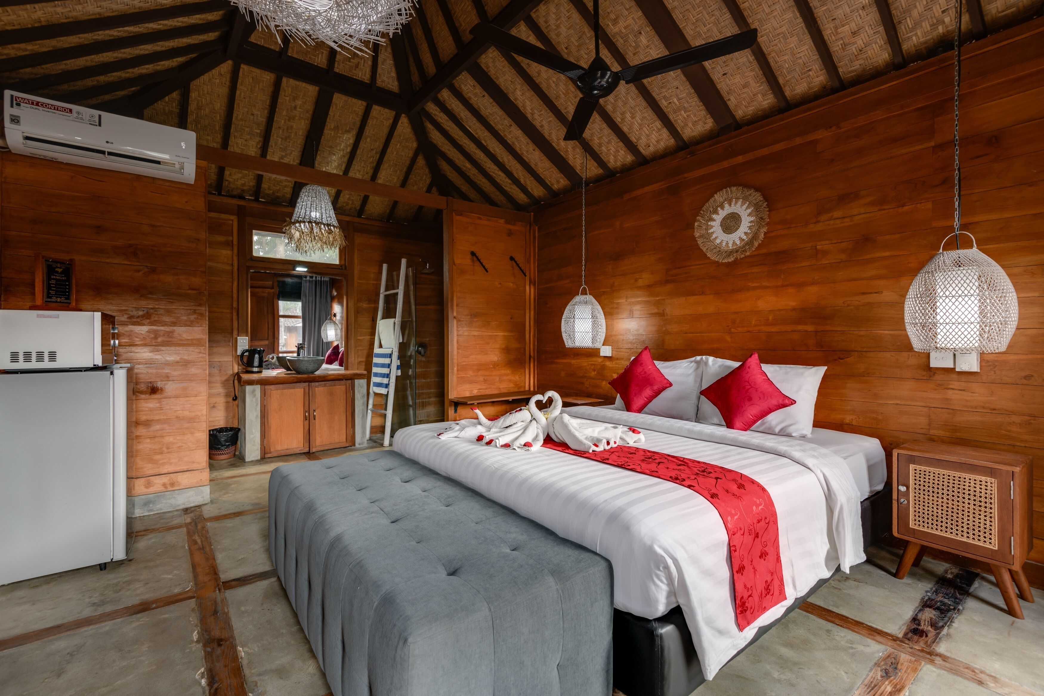 A wooden room with a bed, a small table, a refrigerator, and a ceiling fan.