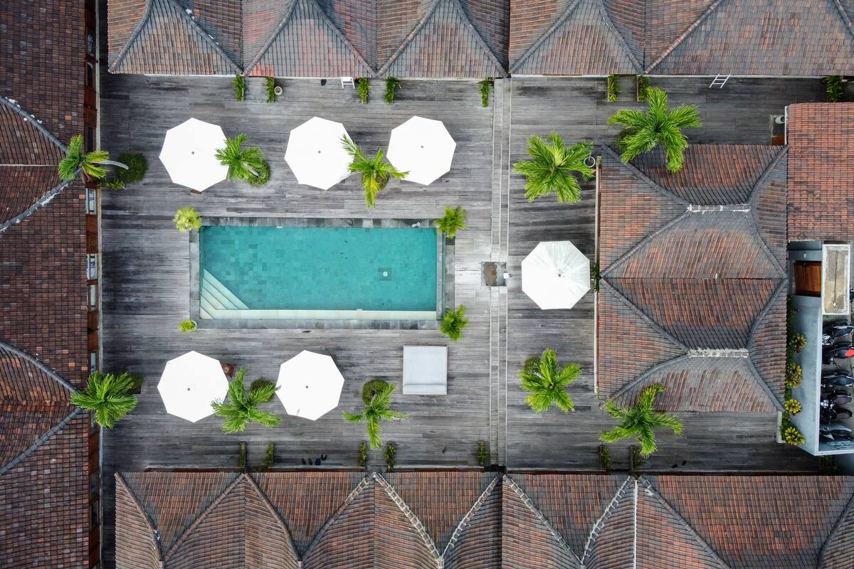 Vista aérea de una zona de piscina en la azotea, con una piscina rectangular, rodeada de sombrillas blancas y palmeras, y un techo con diseño de ladrillo.