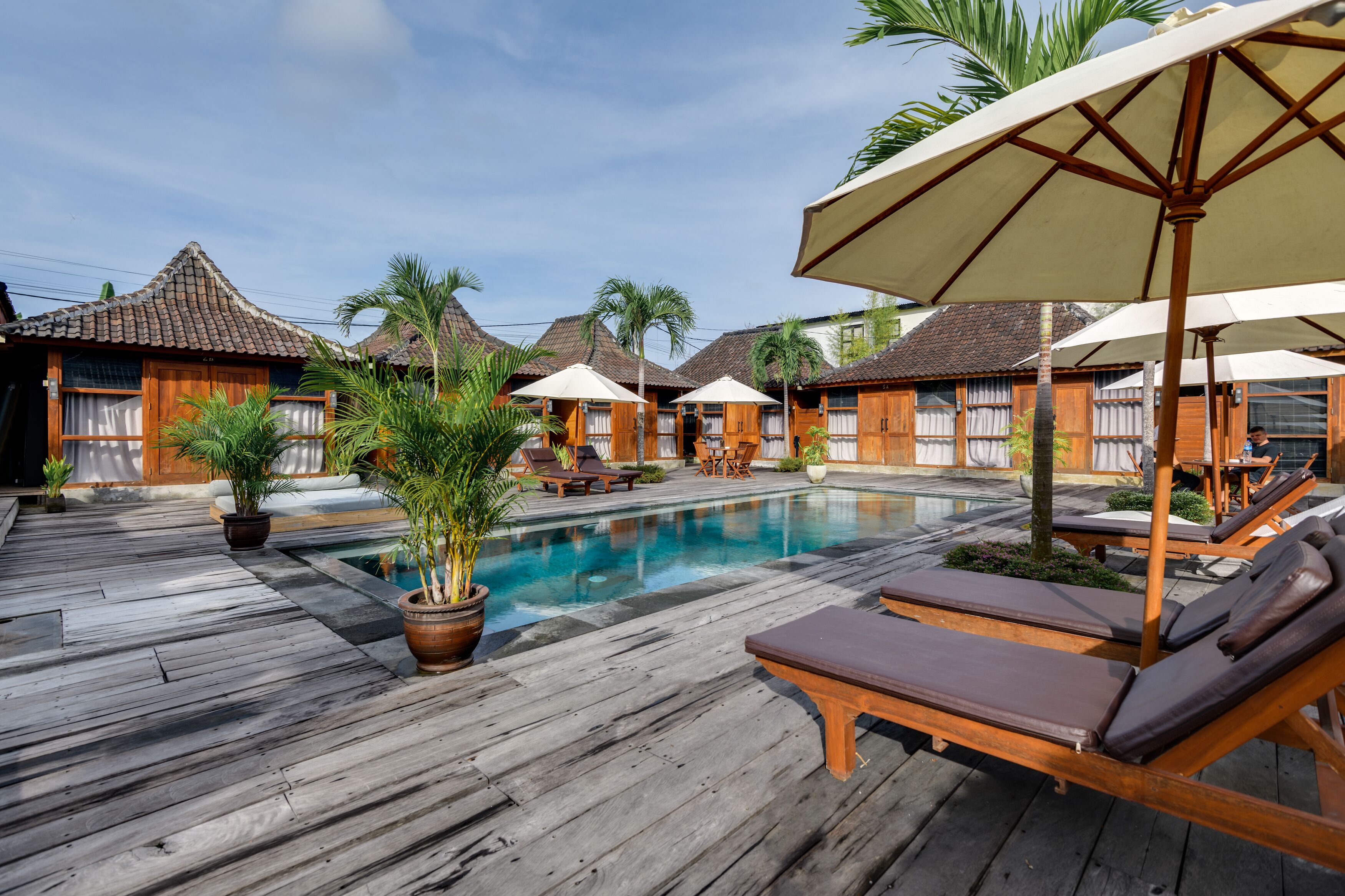 Une terrasse en bois avec des chaises longues et des parasols, une piscine et des bâtiments traditionnels en bois aux toits de chaume.