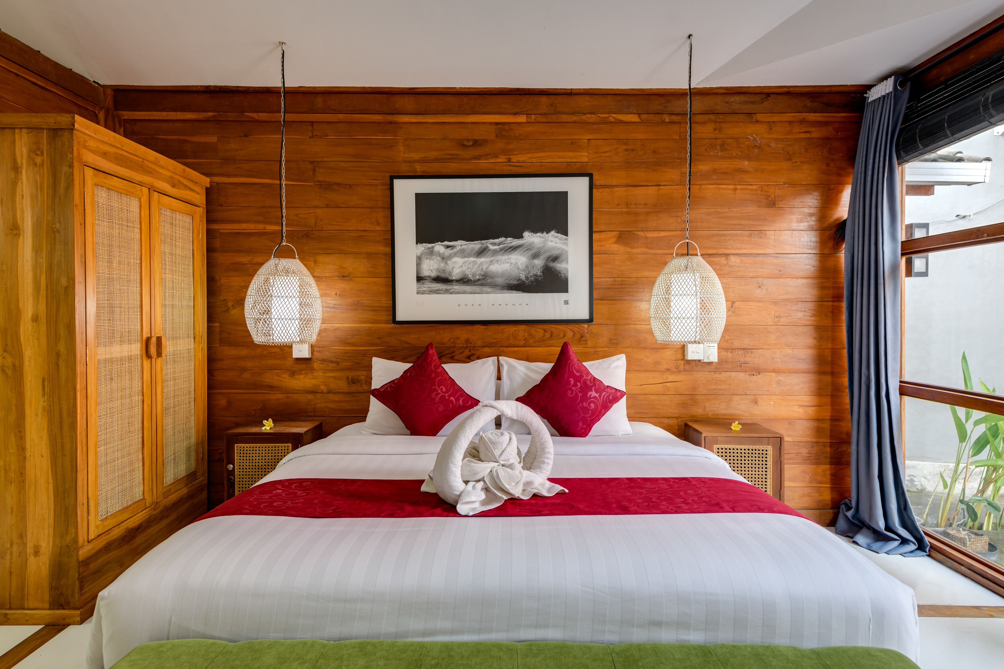 A hotel room with wooden walls, a large bed with white and red bedding, a framed photograph on the wall, and a view of the sea through the window.