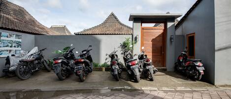 A row of motorcycles parked in a covered area with a tiled floor, a wooden door, and a small building with a tiled roof.