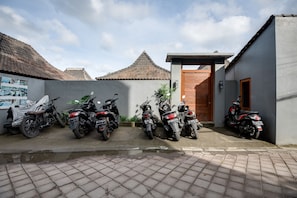 A row of motorcycles parked in a covered area with a tiled floor, a wooden door, and a small building with a tiled roof. - Turtle Villas Canggu (Tibubeneng)