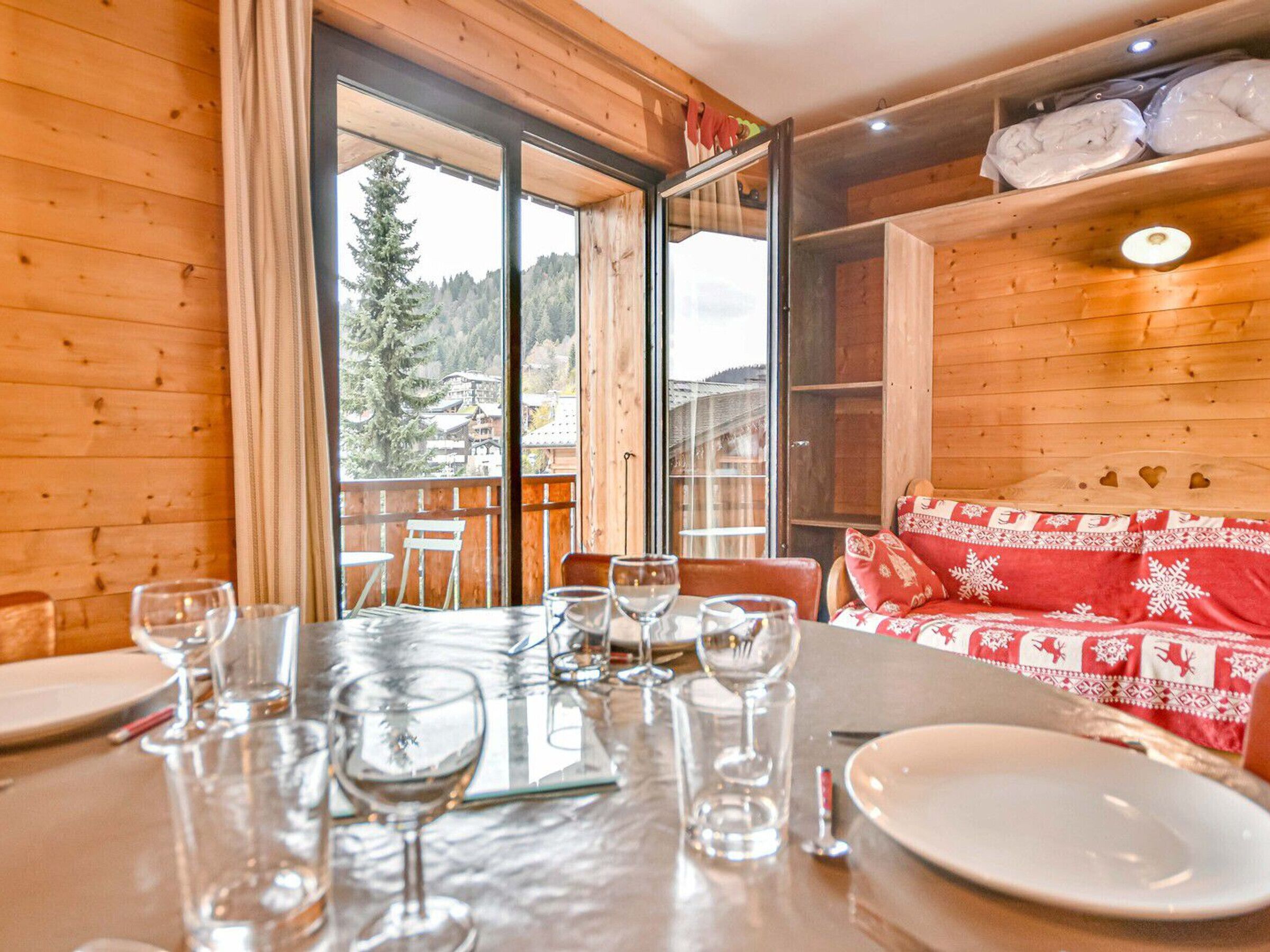 A wooden interior with a dining table set for four, a red and white patterned sofa, and a view of a forested area through large windows.