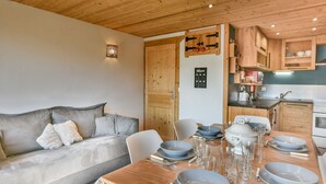 A cozy living room with a sofa, a dining table set for four, and wooden cabinets in the kitchen area. - Cozy 1BR + Alcove, Balcony, Near Slopes and Town, Sleeps 6 (Les Gets)