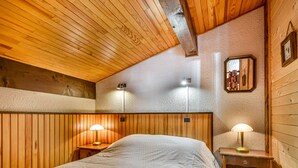 Un dormitorio acogedor con techo de madera, una cama con edredón blanco, dos lámparas de noche y una mesita de noche de madera.