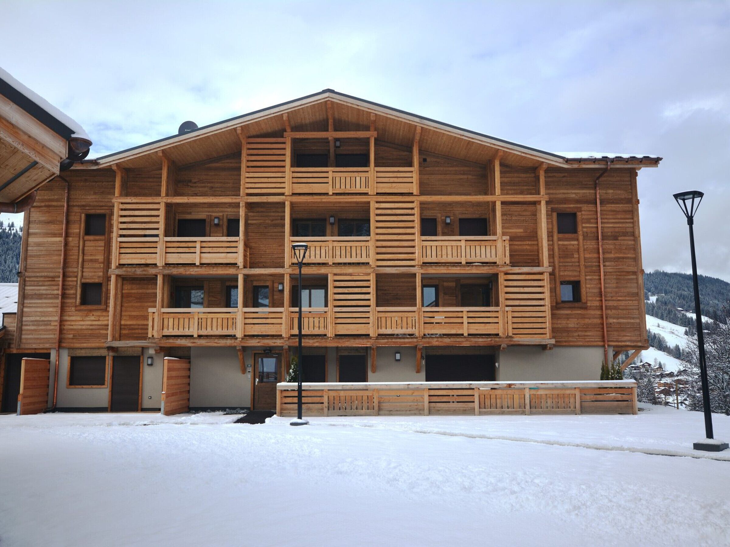 A multi-story wooden building with balconies, a snow-covered ground, and a mountainous backdrop.