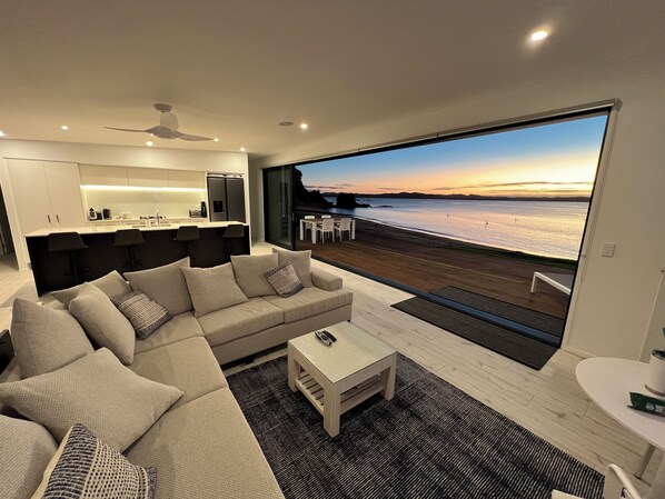 A modern living room with a large sofa, a coffee table, and a view of a beach at sunset.