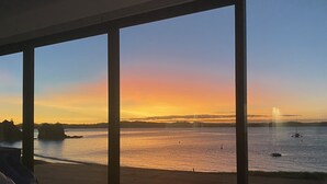 Une pièce dotée de grandes fenêtres donnant sur un paysage où l’on peut admirer le coucher de soleil sur une étendue d’eau parsemée de bateaux.
