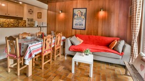 Un salon chaleureux avec un canapé rouge, une table à manger en bois et des chaises. Une petite table blanche est placée devant le canapé. Les murs de la pièce sont en bois et le sol est carrelé.