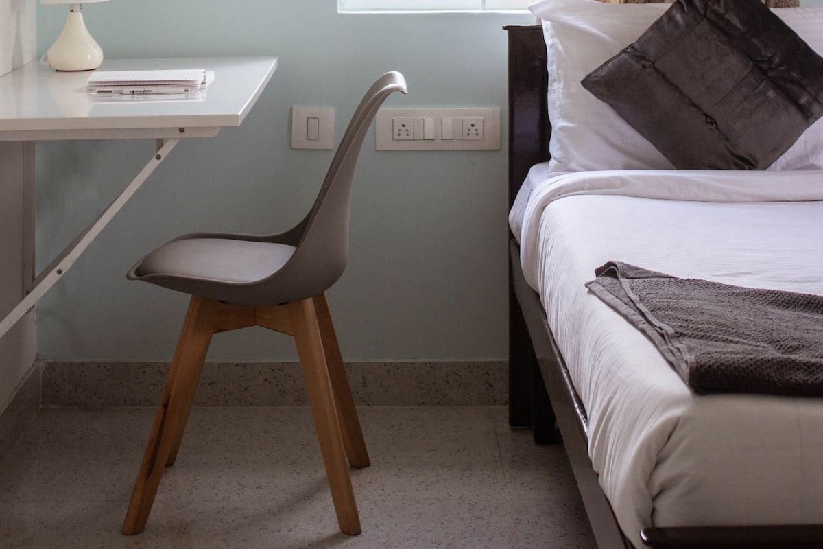 Un dormitorio con cama, escritorio, silla, ventana con cortinas y mesita de noche.