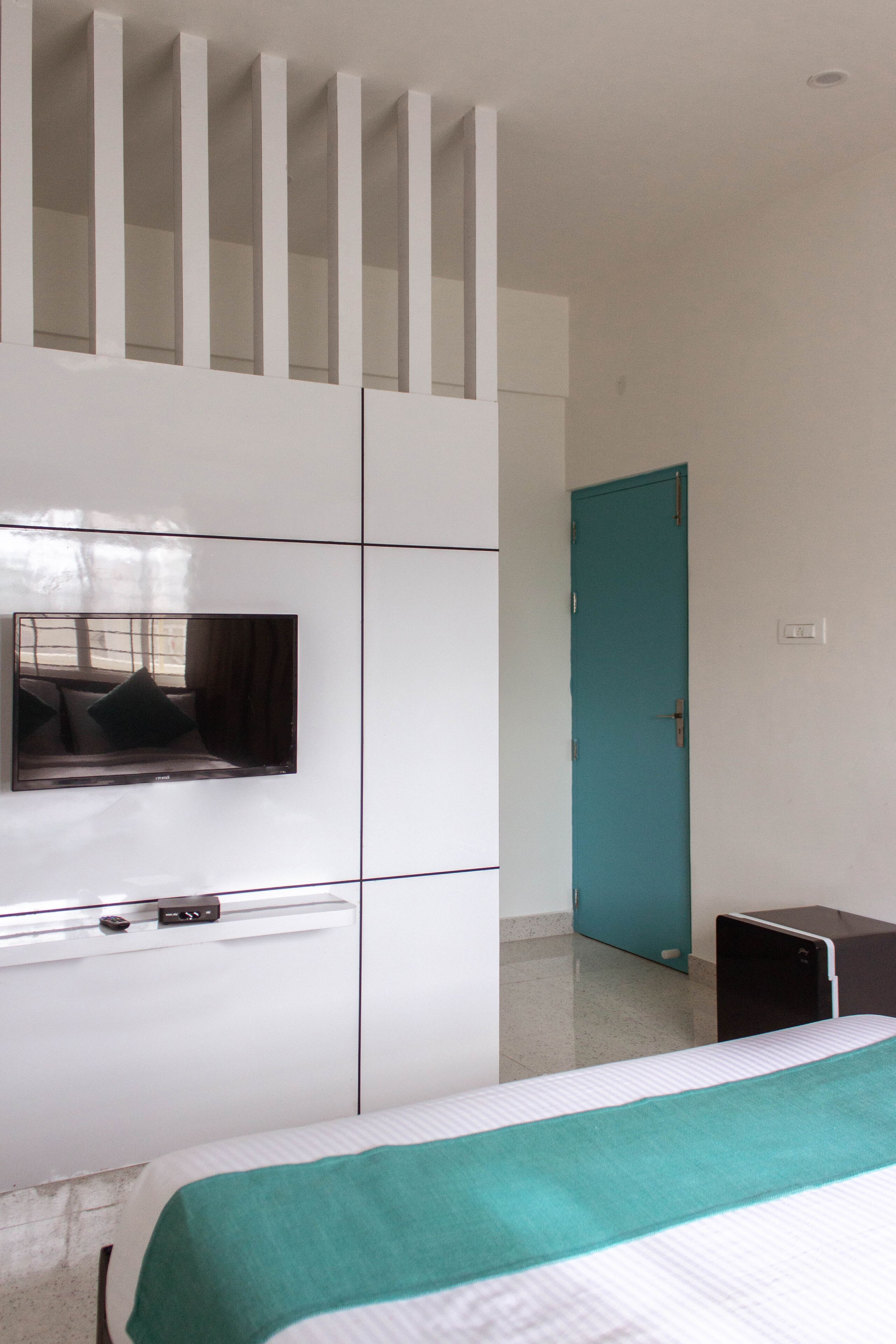 A bedroom with a bed, a TV mounted on a white wardrobe, a teal door, and a small desk.